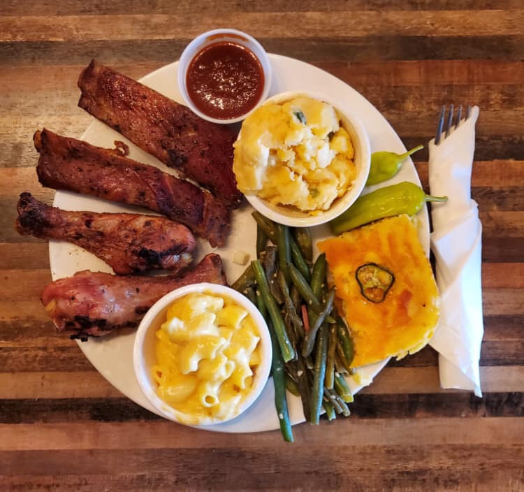 Plate of BBQ with side dishes of potato salad, mac and cheese, green beans, corn bread on a plate with silverware wrapped in a napkin on a wooden table. 